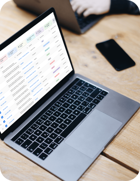 A silver laptop displaying a list interface on a wooden table, with a phone and a second, partially visible laptop in the background.